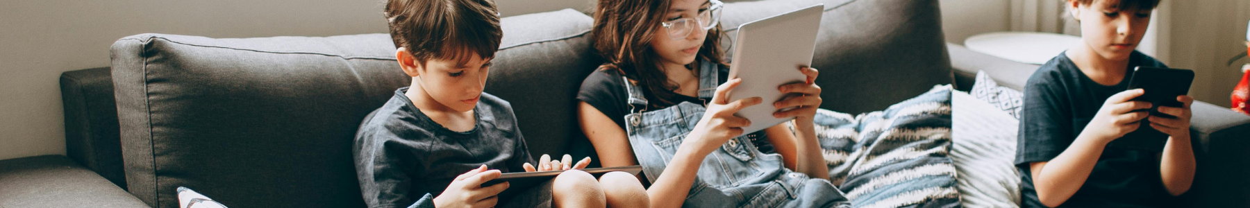 three children sitting on a sofa at home each of them playing on a phone or tablet